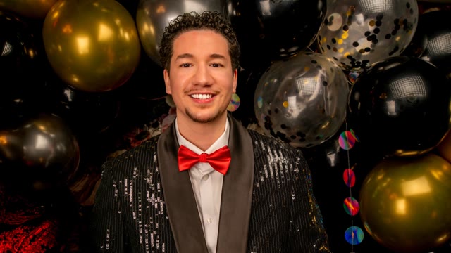 A guest in a sequinned jacket and red bow tie smiles in front of gold and black balloons