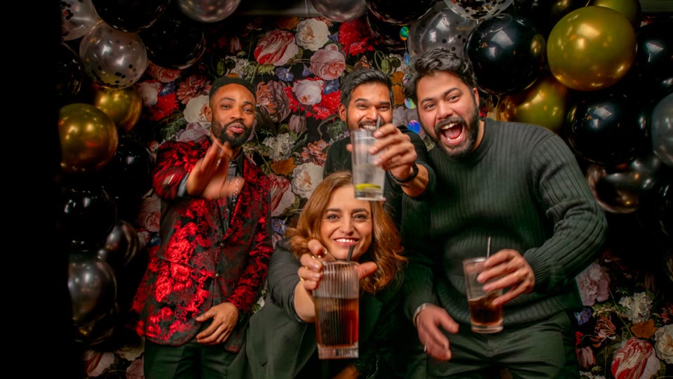 Four colleagues laugh in front of a wall of balloons and flowers and raise their glasses to the camera