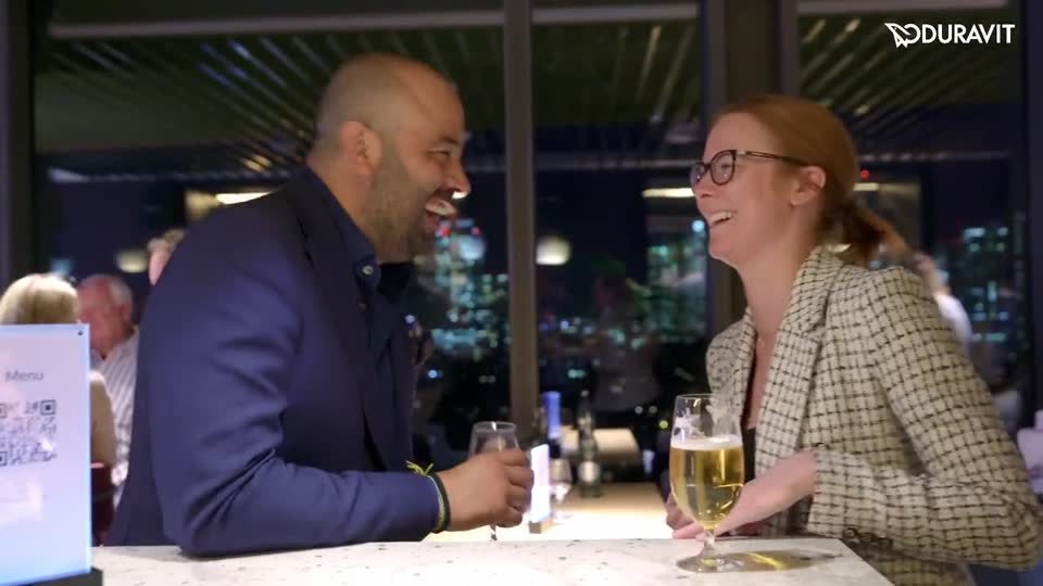 Two guests laughing at the bar of a rooftop restaurant, Frankfurt lights behind them