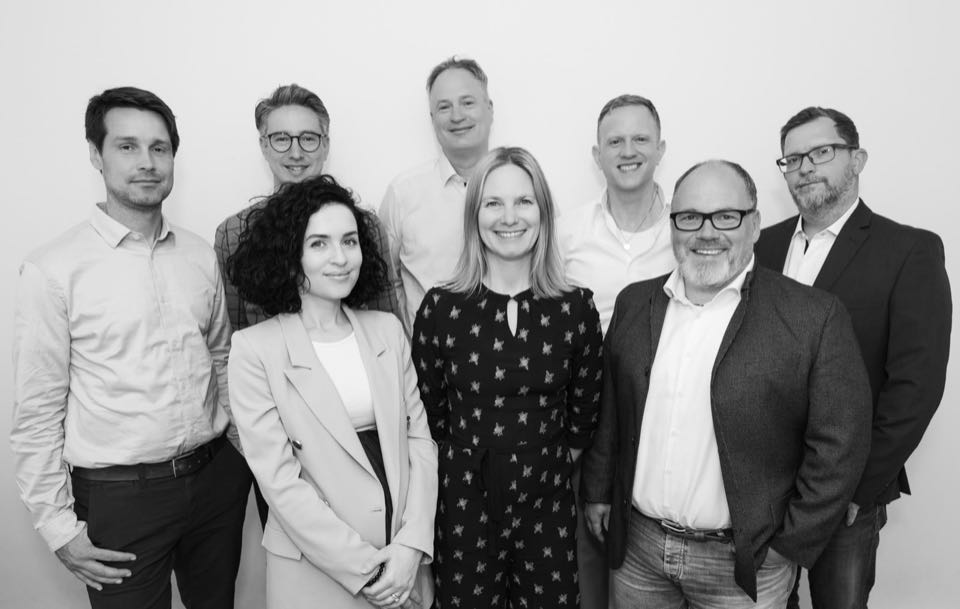 Schwarzweißes Teamfoto: acht Kolleginnen und Kollegen in Businesskleidung vor einer weißen Wand