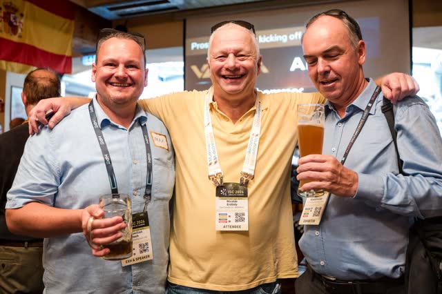 Drei Gäste mit Namensbändern und Bier lachen Arm in Arm beim Abendempfang