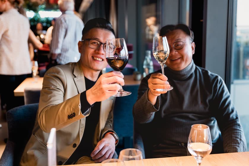 Zwei Gäste heben am Restauranttisch ihre Weingläser in die Kamera